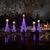 The image shows a festive night scene with people admiring several illuminated, cone-shaped light displays resembling Christmas trees, set against a backdrop of trees and buildings adorned with holiday decorations.