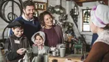 A happy family is engaging with a person in historical attire at a colonial workshop, surrounded by metalwork items.