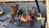 A group of jovial people pose for a photo on a sailboat during a sunny day, with the backdrop of tranquil waters and a clear sky.