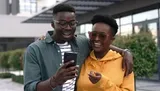 Two people are smiling and looking at a smartphone together with a backdrop of urban architecture.