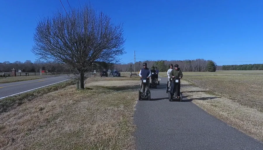 A group of people is riding Segways on a paved path in a scenic area with trees and a clear blue sky.