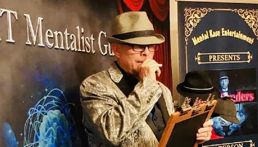 A performer in a glittery jacket and fedora holds a clipboard and appears deep in thought at an event presented by Mentalist G.