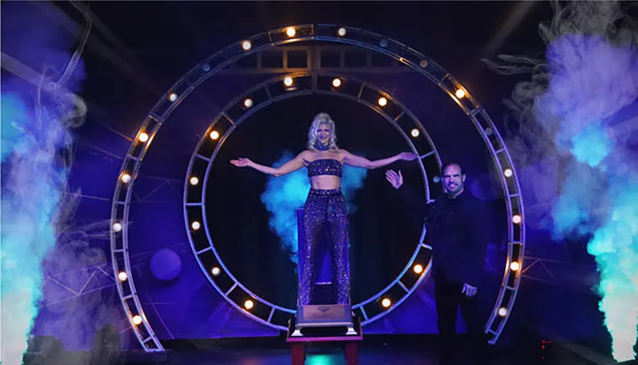 A magician poses next to an assistant standing inside a lit circular portal surrounded by theatrical smoke on stage.