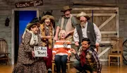 A group of costumed actors is posing enthusiastically on a theater set that evokes a wild west theme, with one actor holding a sign that reads 