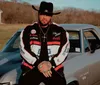 A man wearing a black hat and a racing jacket leans on a vintage muscle car in a rural setting