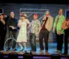 A group of eight people in colorful attire is joyfully posing on a stage with one person holding a guitar and another with a unicycle