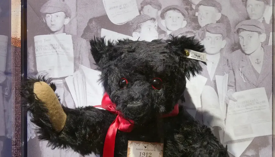 A black teddy bear with a red ribbon and tag is displayed against a vintage photograph background.