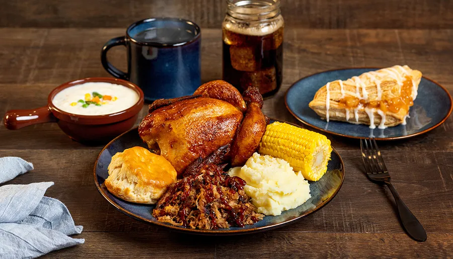 The image features a hearty meal with roasted chicken, corn on the cob, mashed potatoes, biscuits, pulled pork, a bowl of soup, a mug of beverage, and a slice of pie, all presented on a wooden table.