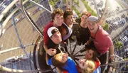 A group of joyful friends takes a selfie from a high vantage point on a sunny day, with their faces close together and a parking lot visible below.