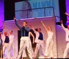 Performers in sparkling costumes are singing and dancing on stage during a production that appears to be themed around the musical Mamma Mia based on the screen projection and attire