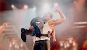 A wrestler is performing a victory pose in the ring, showcasing his muscles, while holding another wrestler on his shoulder.