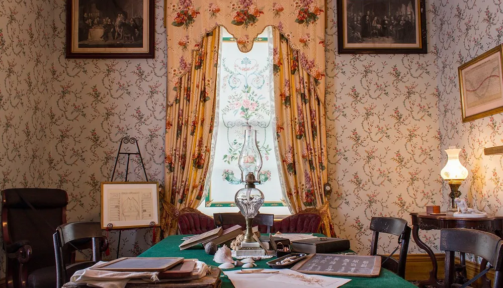 The image shows a vintage room with floral wallpaper and drapery furnished with a table laden with books documents a lamp and chairs evoking a historical atmosphere