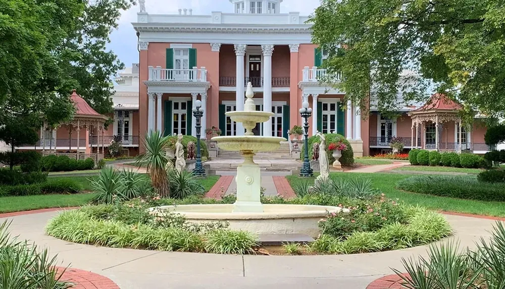 The image features an elegant pink-hued mansion with white columns and balconies flanked by two smaller structures set behind a manicured garden with a classic water fountain as the centerpiece