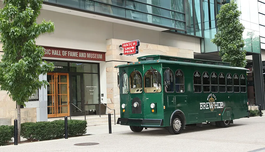 A vintage-style green trolley bus labeled Music City BrewHop is parked in front of a building with a sign that reads Music Hall of Fame and Museum.