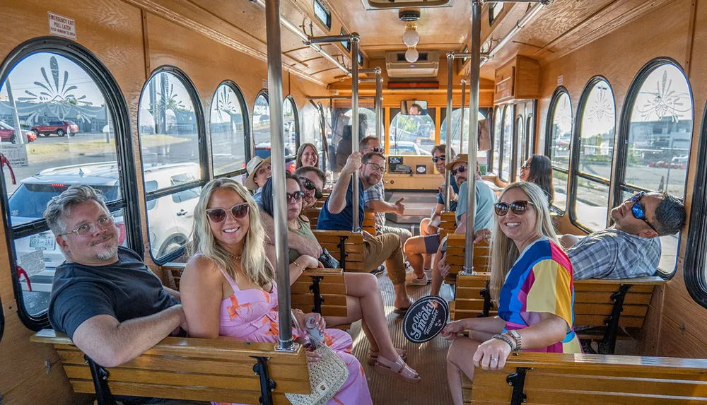 A group of cheerful people are seated inside a vintage-looking trolley car with large windows enjoying their ride under a bright sunny sky