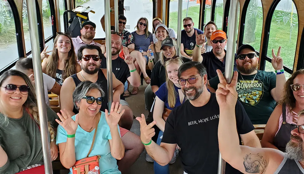 A group of cheerful people are posing for a photo while riding on a bus or tram with many of them smiling and making hand gestures to the camera