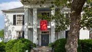 The image shows a two-story white house with black shutters, a red entrance door, and a red banner hanging from the second-floor balcony, all framed by green foliage and a prominent tree.