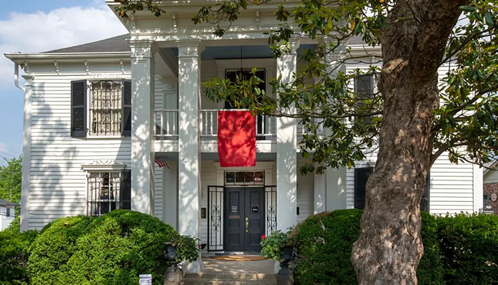 The image shows a two-story white house with black shutters a red entrance door and a red banner hanging from the second-floor balcony all framed by green foliage and a prominent tree