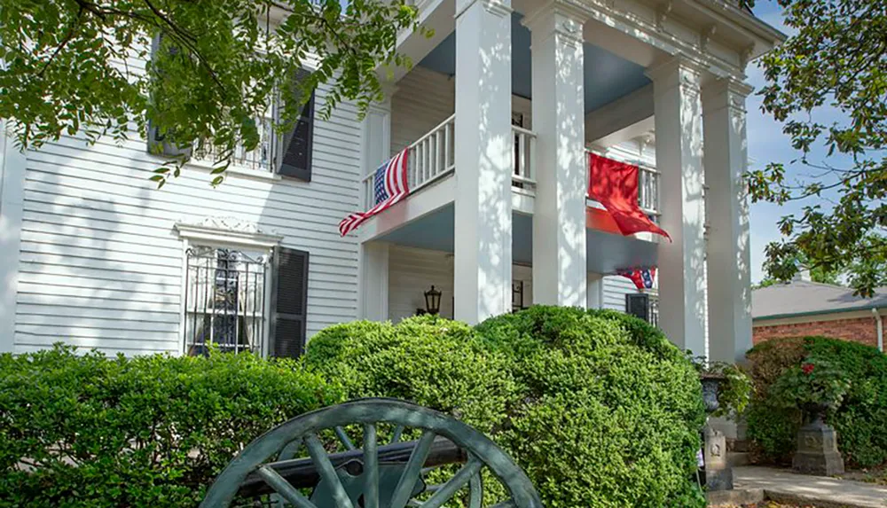 The image shows the entrance of a classic two-story American home with a white facade front porch columns American flag and red flag all framed by lush greenery and a vintage wheel decoration in the foreground