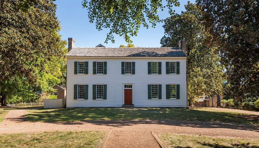 The image features a two-story white colonial house with green shutters and a red door surrounded by trees and a well-kept lawn under a clear blue sky