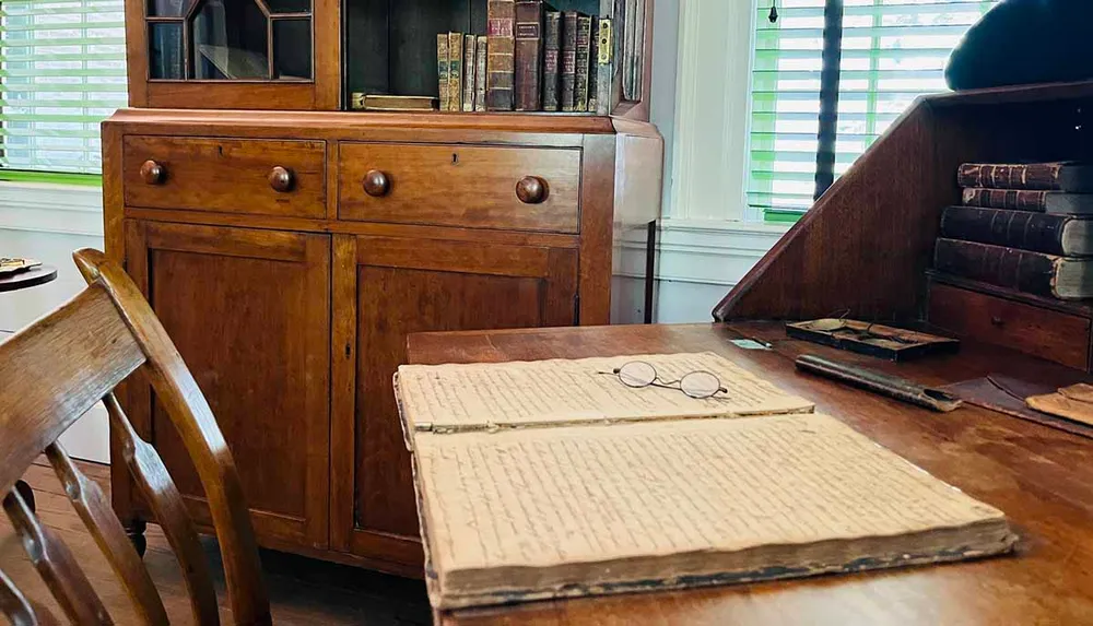 The image depicts a vintage wooden writing desk with an open ledger or book a pair of round glasses and several old books evoking a historical and scholarly atmosphere