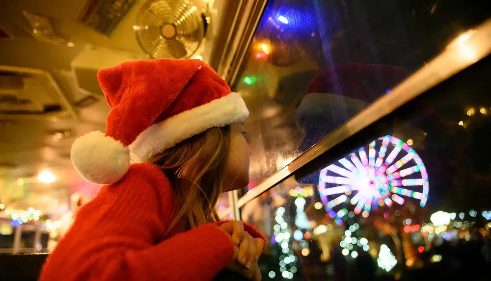 A child in a Santa hat gazes out a window at night captivated by the colorful lights of a Ferris wheel and holiday decorations