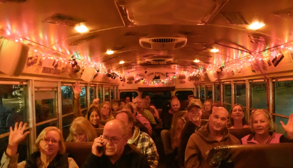 A group of cheerful people is sitting inside a bus adorned with festive lights smiling and waving at the camera