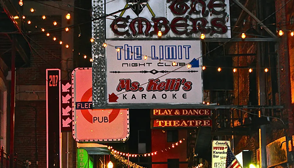 The image shows a vibrant street scene at dusk with various signs advertising establishments such as a night club karaoke a pub and a play  dance theatre all adorned with twinkling string lights