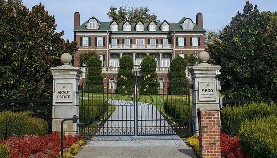 The image shows a grand, brick estate with a gated entrance, surrounded by trimmed hedges and trees.