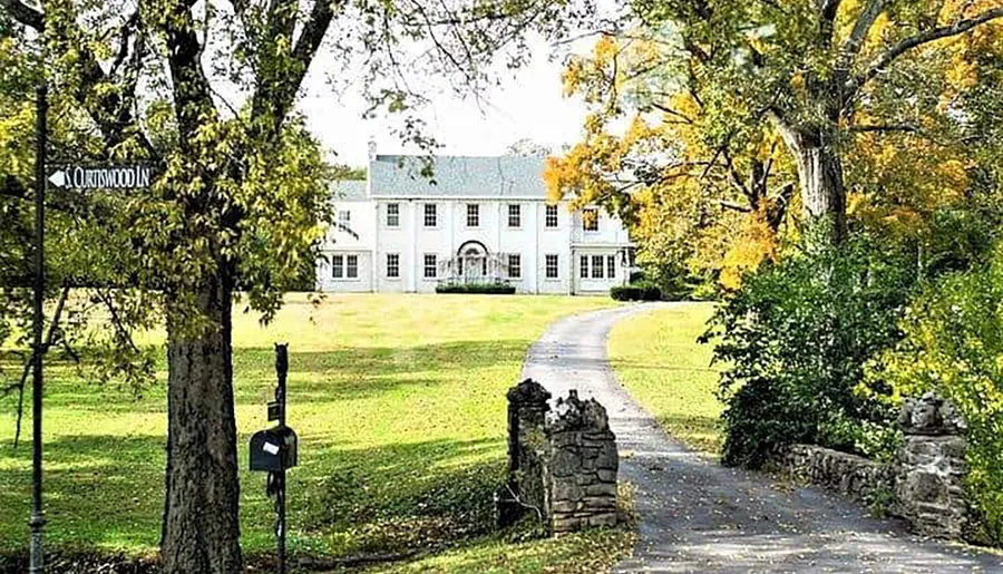 A picturesque lane leads to a large white house surrounded by trees with autumn foliage.