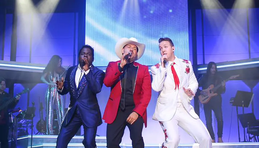 Three male performers are singing passionately on stage with a band visible in the background, illuminated by bright stage lights.