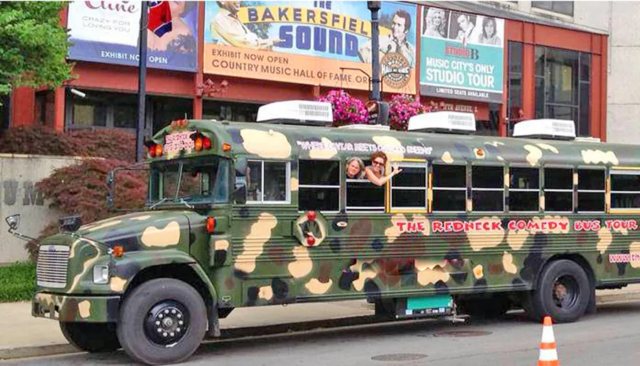 A camouflage-painted bus with the text The Redneck Comedy Bus Tour parked in an urban area, with two people peeking out from the windows and waving.