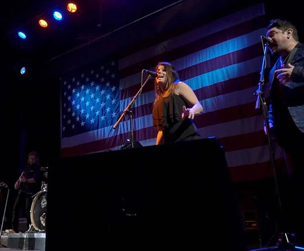 A band performs on stage with a large American flag as the backdrop with a singer engaging passionately with the microphone and another band member playing the guitar