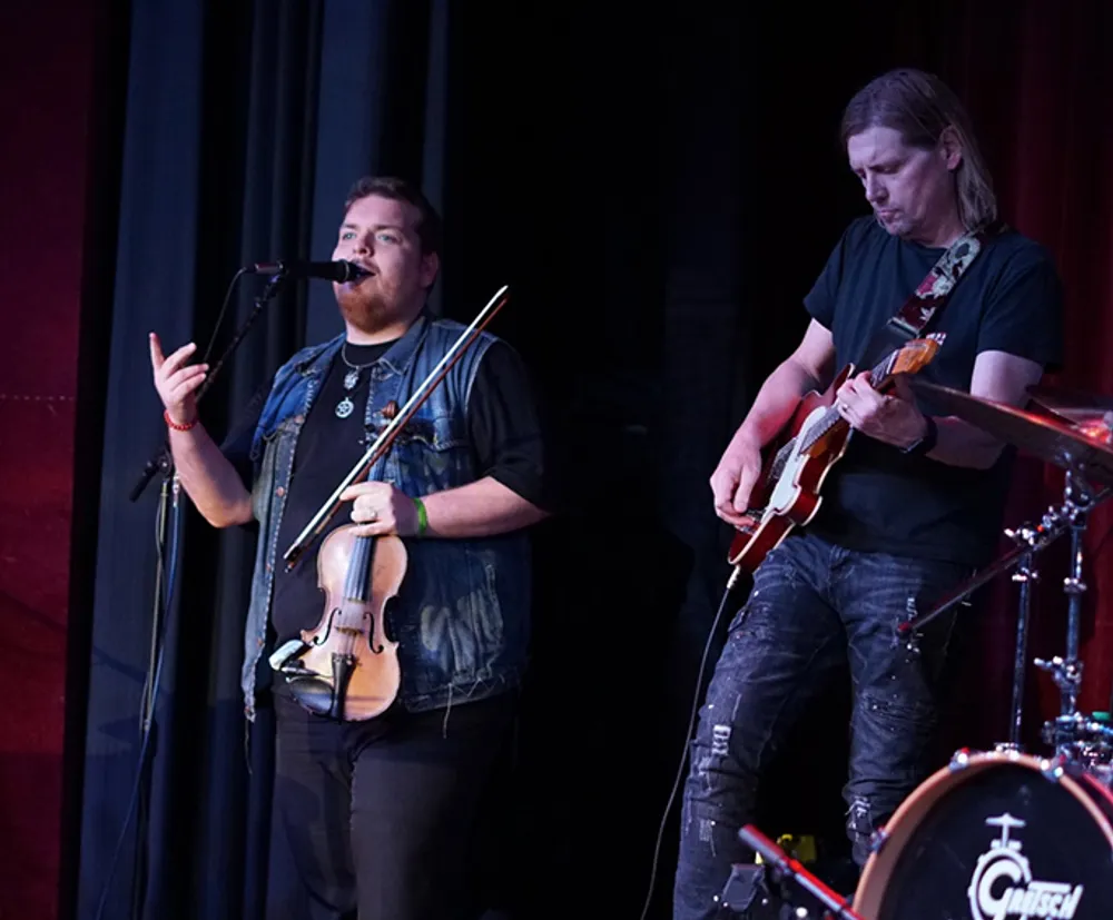 Two musicians are performing on stage with one playing the violin and singing into a microphone and the other playing the electric guitar