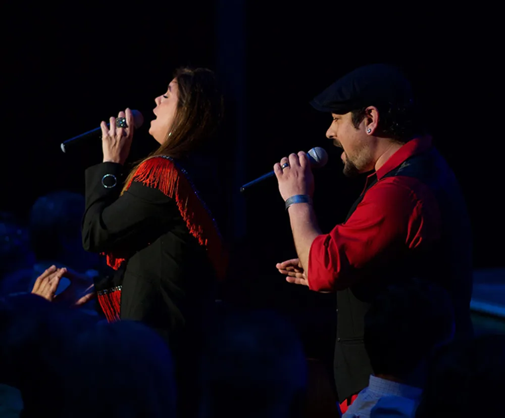 A man and a woman are passionately singing into microphones on stage in front of an audience