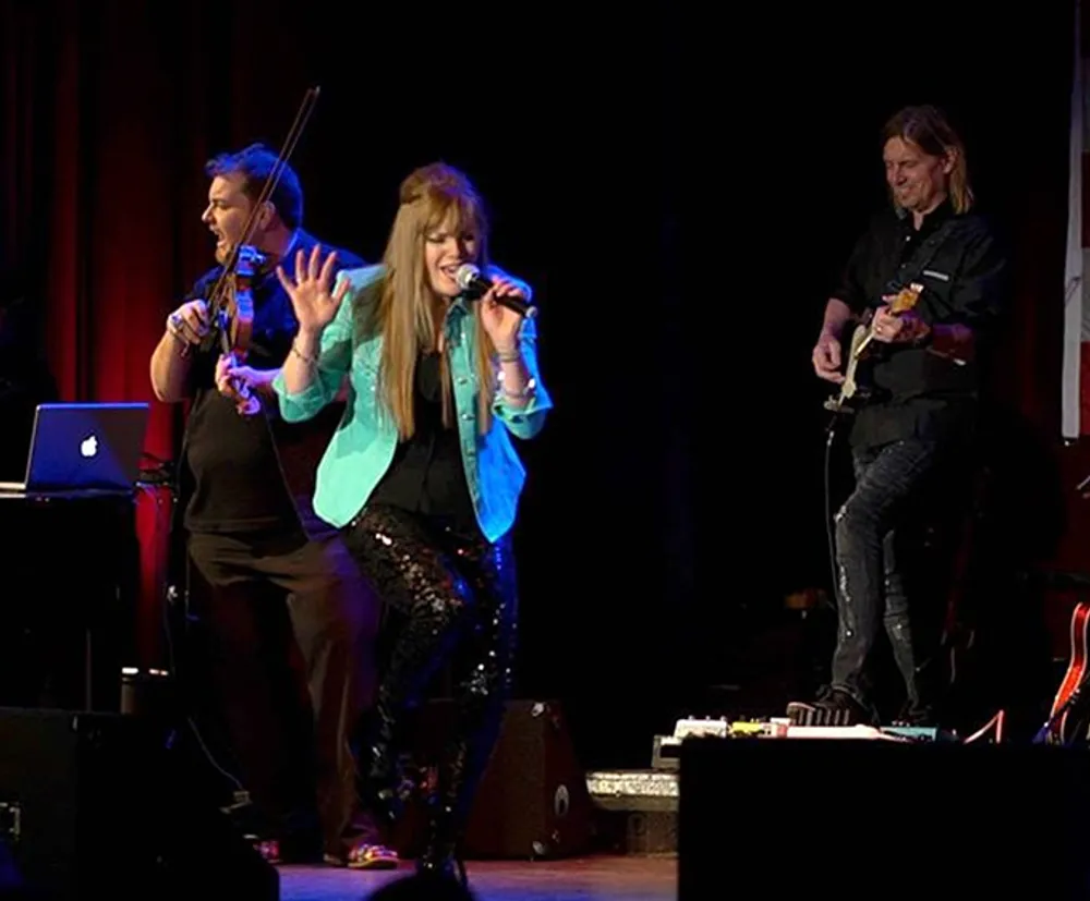 A female vocalist passionately performs on stage accompanied by a violinist and a guitarist