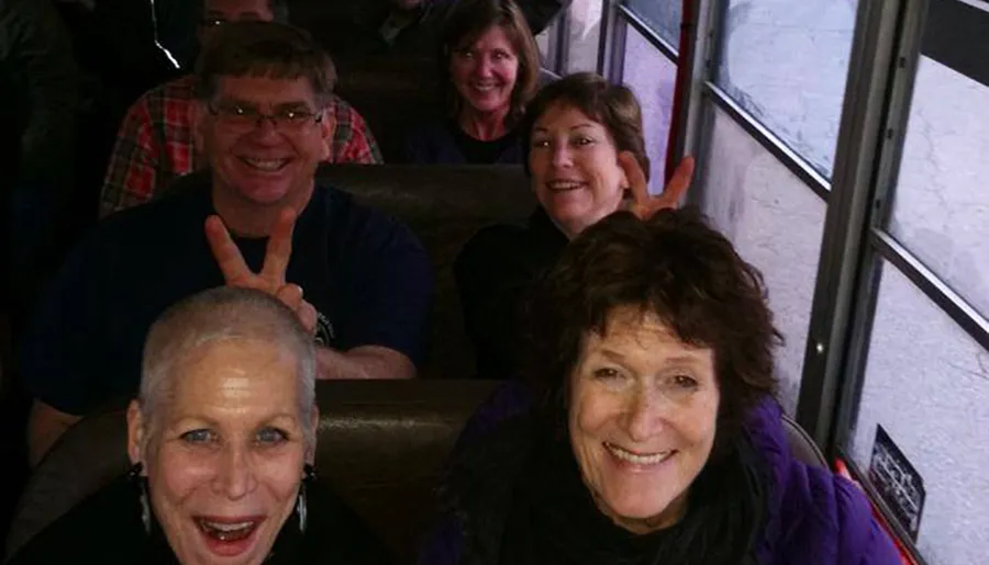 A group of smiling individuals are enjoying a moment together on a bus, with one person playfully making a peace sign behind another's head.