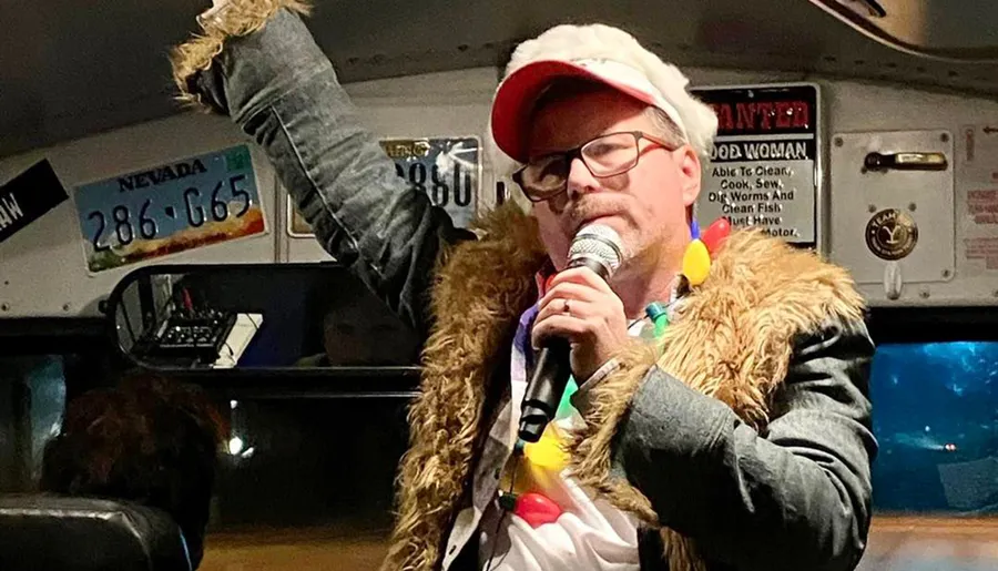 A person wearing a Santa hat and a jacket with fur trim holds a microphone and is gesturing animatedly in a space decorated with novelty lights and what appears to be a license plate.