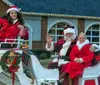 Three individuals dressed in festive holiday attire are sitting in a white horse-drawn sleigh adorned with Christmas decorations waving and smiling in front of a building with holiday lights