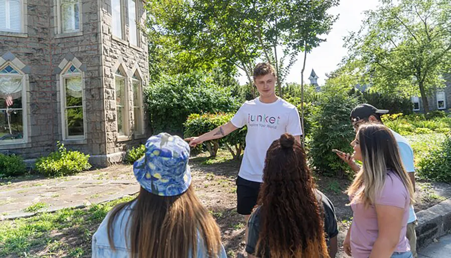 A person is standing and pointing something out to a group of attentive listeners outdoors near a stone building.