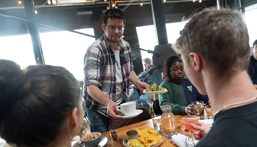 A person is serving food at a busy dining table where several other people are seated with drinks and plates in front of them.