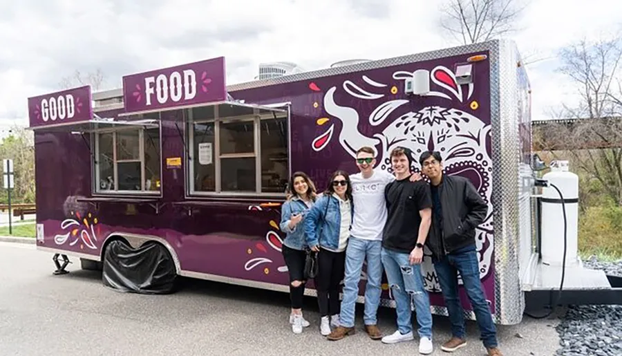 A group of five people are smiling and posing in front of a purple food truck with the words GOOD FOOD written on it.