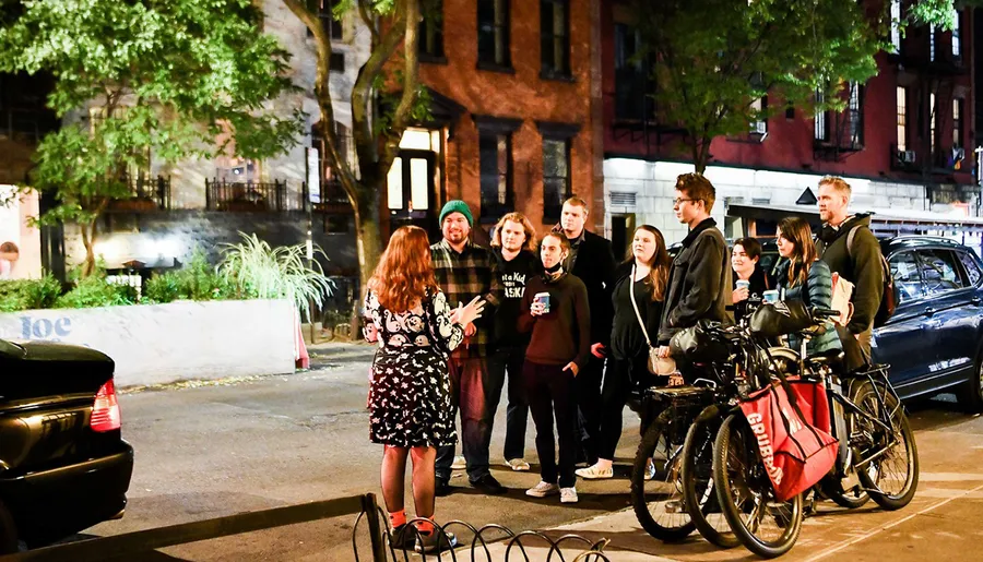 A group of people is engaging in a conversation on a city street at night, with some standing by bicycles and a car parked nearby.