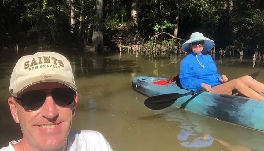 A man in a cap is taking a selfie with a woman in a blue shirt and hat sitting in a kayak on a river surrounded by trees.