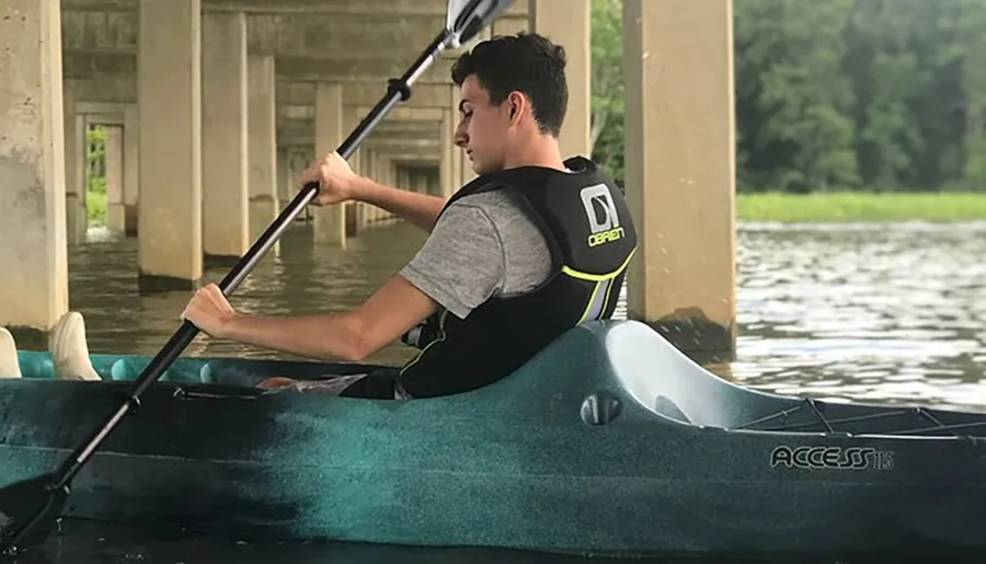 A person is kayaking under a bridge on a calm body of water, wearing a life vest and actively paddling.