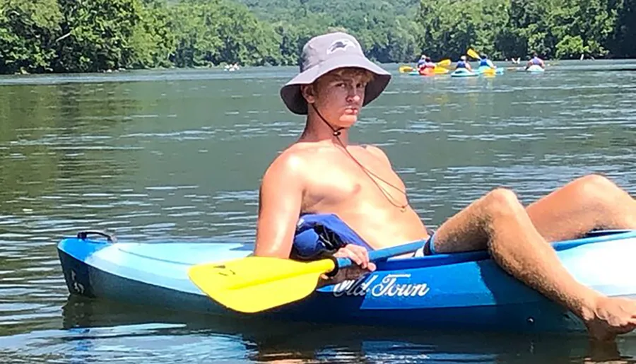 A person lounges on a blue kayak in calm waters wearing a wide-brimmed hat and holding a yellow paddle.