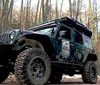 A rugged off-road vehicle equipped with roof-mounted gear is parked in a woodland area near a simple wooden structure with a solar panel on its roof