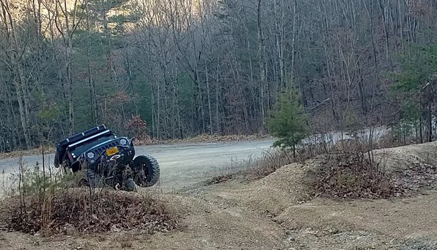 A modified off-road vehicle is traversing a rough, hilly terrain with a background of leafless trees.