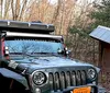 A rugged off-road vehicle equipped with roof-mounted gear is parked in a woodland area near a simple wooden structure with a solar panel on its roof