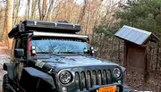 A rugged, off-road vehicle equipped with roof-mounted gear is parked in a woodland area near a simple wooden structure with a solar panel on its roof.